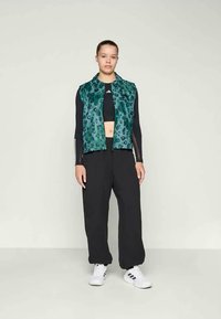 Žena stoji odjevena u teal prsluk sa leopard uzorkom preko crnog dugog rukava kropske majice, labavim crnim hlačama i bijelim tenisicama s crnim prugama.