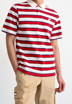 Mann trägt ein weißes Polohemd mit roten und blauen horizontalen Streifen und beige Cargohosen, steht mit entspannten Händen.