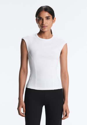 Sötét hajú nő fehér, ujjatlan, testhezálló felsőben és fekete nadrágban, egyszínű, világos háttér előtt állva.