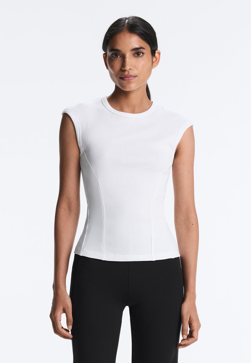 Sötét hajú nő fehér, ujjatlan, testhezálló felsőben és fekete nadrágban, egyszínű, világos háttér előtt állva.