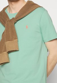 Mentolově zelené tričko s krátkým rukávem a malým kontrastním oranžovým logem. Hnědý texturovaný pletený svetr je přehozený přes ramena.