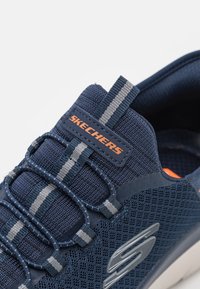 Mörkblå sneakers med ovandel av mesh-textil, kontrasterande grå ränder, elastiskt snörsystem och oranga logodetaljer på tungan.