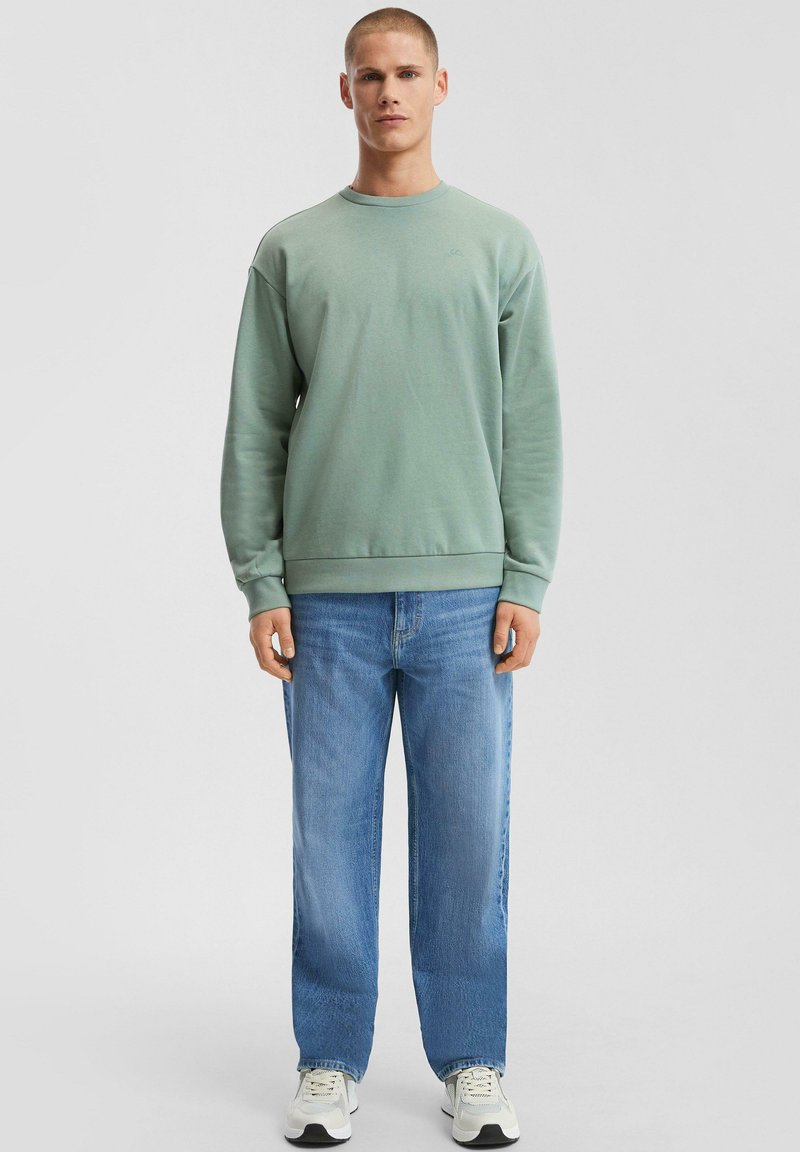 Giovane in piedi, indossa una felpa verde salvia, jeans azzurri chiari e sneakers bianche su uno sfondo grigio chiaro uniforme.