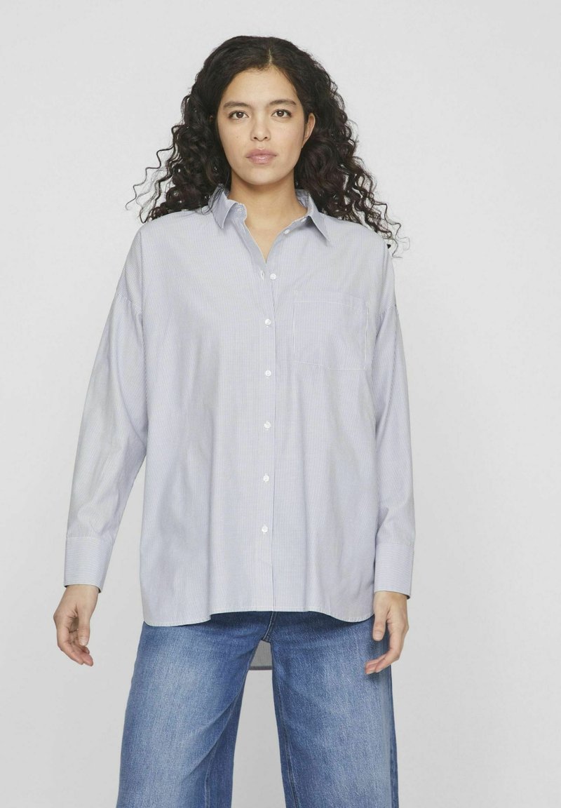 Camisa de mangas largas a rayas en azul claro y blanco con cuello abotonado, bolsillo frontal y corte relajado. Combinada con pantalones de mezclilla holgados.