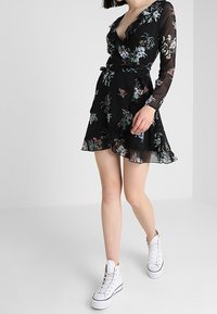 Schwarzes Blumenkleid mit Wickeldesign, langen Ärmeln und Rüschen. Leichter Stoff, mit einem Bindegürtel. Kombiniert mit weißen Sneakers.