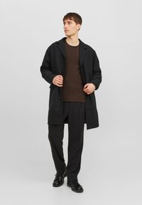 Svart överrock i tungt tyg med snedkrage, framknappar och avslappnad passform, bärs över en brun texturerad skjorta och svarta byxor.