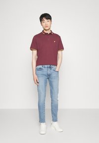Burgundy pikétröja med gul trim, matchad med ljusblå skinnyjeans och vita sneakers, med en liten logotyp på bröstet.