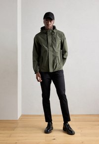 Olive green waterproof jacket with a hood, zip and snap closures, side pockets, and black fitted pants with black shoes, on a wooden floor.