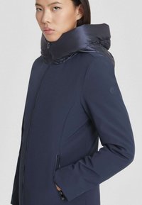 Doudoune marine avec col haut et accents brillants. Conçue en matériau doux, dotée de poches latérales zippées et d'un design épuré.