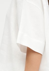 Posnetek v bližini roke osebe, ki nosi oblačilo iz bele teksturirane tkanine s kratkimi, ohlapnimi in širokimi rokavi.