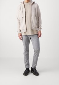 Felpa grigia chiara con zip sopra una maglietta beige, abbinata a pantaloni grigi a quadri e scarpe stringate nere con suola spessa.