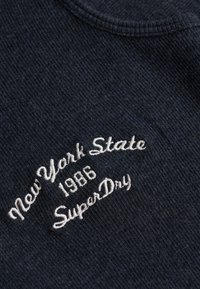 Tissu côtelé sombre avec un texte blanc brodé indiquant "New York State 1986 SuperDry" en disposition incurvée.
