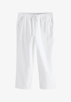 Junge, der in weißen Hosen, schwarzem kurzärmeligen Hemd mit Knöpfen und braunen Sandalen vor einem schlichten beigen Hintergrund steht.