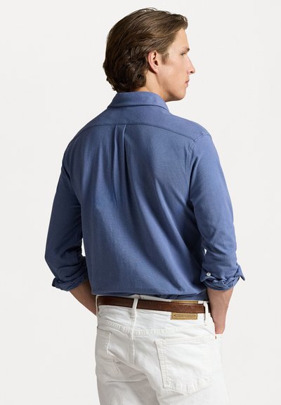 Camicia blu a maniche lunghe con maniche arrotolate, taglio sartoriale e piega sul retro. Abbinata a pantaloni bianchi e una cintura marrone.