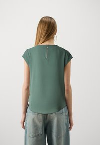 Grünes Kurzarmshirt mit rundem Ausschnitt und einem Reißverschlussdetails im Rücken. Getragen mit hellblauen Jeans aus Denim mit Gesäßtaschen.
