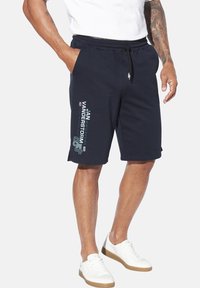 Marineblaue Baumwollshorts mit elastischem Bund und Kordelzug, ausgestattet mit einem weißen und grünen Drucklogo auf der linken Seite, kombiniert mit weißen Turnschuhen.