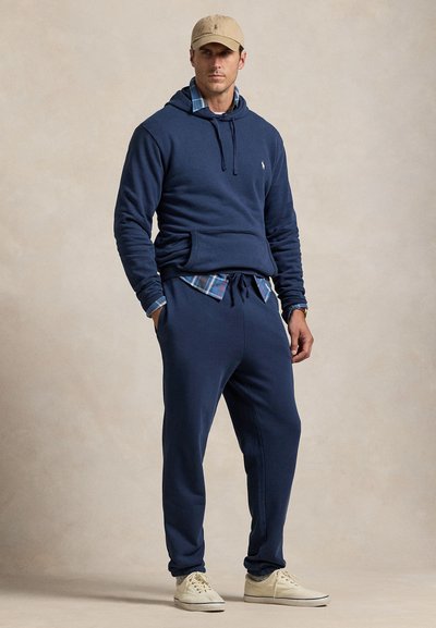 Marineblå hættetrøje med en forlomme, kombineret med matchende joggingbukser og en ternet skjorte under. Har en kasket og hvide sneakers.