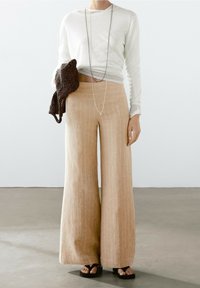 Beige linnen broek met wijde pijpen en verticale strepen, gecombineerd met een licht, lang mouwtopje en een getextureerde donkere tas. Eenvoudige zilveren ketting als accent.