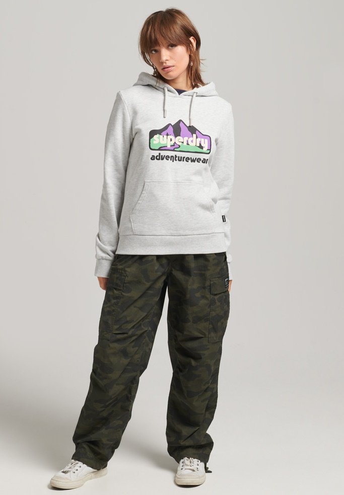 Felpa grigia con tasca frontale e logo viola/verde, abbinata a pantaloni cargo mimetici larghi. Sneaker bianche completano l'outfit.