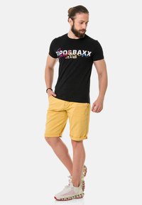 Schwarzes Baumwoll-T-Shirt mit buntem Logo-Text, kombiniert mit gelben Denim-Shorts mit umgeschlagenen Säumen und roten Akzenten. Weiße Sneakers mit strukturiertem Design.