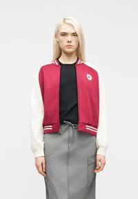 Ξανθιά γυναίκα που φοράει κόκκινο και λευκό varsity μπουφάν πάνω από μαύρο τοπ και γκρι φούστα cargo, στέκεται μπροστά σε απλό λευκό φόντο.