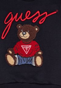 Miś pluszowy ubrany w czerwony sweter Guess z logo oraz niebieskie spodnie, wyszyte na ciemnej tkaninie pod czerwonym napisem Guess.