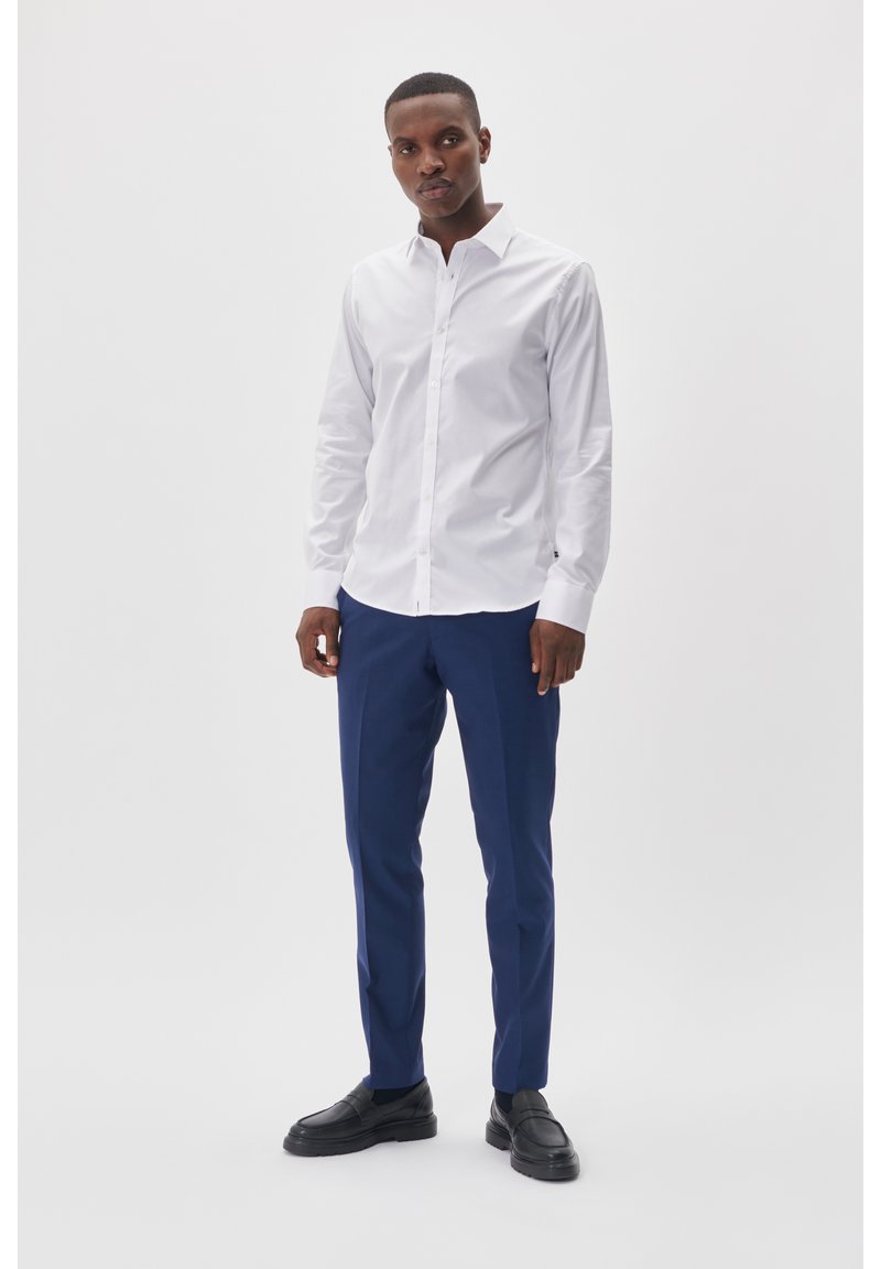 Witte blouse met knoopsluiting, gecombineerd met blauwe broek. Model draagt zwarte instappers. Gladde stof en nauwsluitende pasvorm.