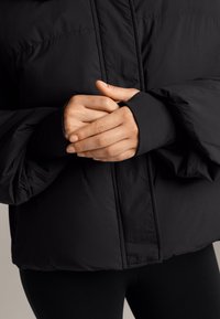 Čierna prešívaná bunda s vysokým golierom, hladkou textúrou a rebrovanými manžetami, nosená s čiernymi legínami, ruky zložené pred telom.