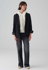 Zwarte blazer over een witte hoodie, met grijze flared jeans en casual schoenen. De outfit heeft een mix van texturen en neutrale kleuren.