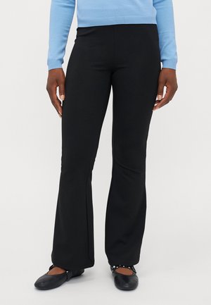 Person trägt schwarze Schlaghosen, schwarze Mary Jane Ballerinas mit Nieten und ein hellblaues Langarmshirt, steht vor weißem Hintergrund.