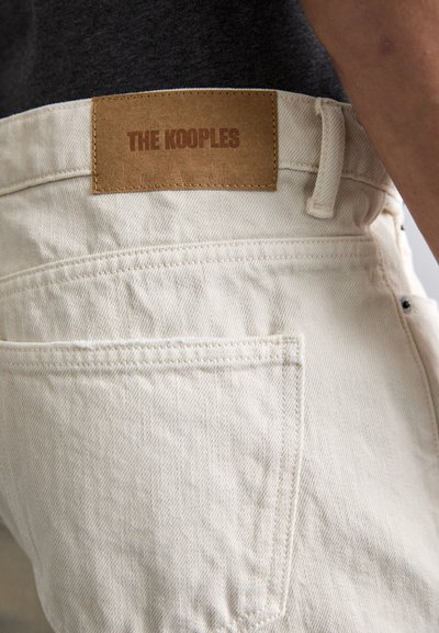 Beige denim jeans med en teksturert overflate, med et brunt lærdeksel merket "THE KOOPLES" på livet. Vanlig fem-lomme design.
