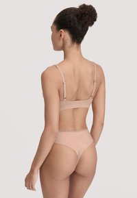Femme aux cheveux bouclés en chignon, portant un soutien-gorge couleur nude et une culotte taille haute assortie avec dentelle, de dos sur fond uni.