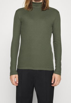 Persona che indossa un maglione a collo alto a costine verde oliva e pantaloni neri, in piedi davanti a uno sfondo chiaro uniforme.