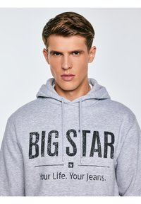 Noor mees lühikeste pruunide juustega, kannab heledat halli kapuutsiga pusa, mille eespool on trükitud "BIG STAR Your Life. Your Jeans."