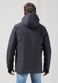 Sort vandtæt jakke med hætte, der har et strømlinet design, elastiske manchetter og en glat overflade, båret over lyseblå jeans.