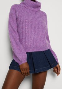 Femme portant un pull à col roulé violet côtelé et une jupe plissée courte bleu marine, debout les mains détendues le long du corps.