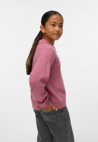 Ung flicka i en rosa tröja och mörkgrå jeans, står på sidan med händerna i fickorna, ser framåt med ett lätt leende.