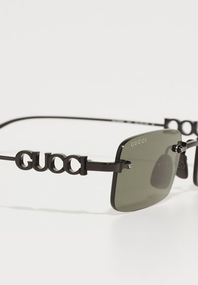 Čierne obdĺžnikové slnečné okuliare so zelenými sklami, ktoré majú elegantný dizajn a výrazné logo "Gucci" na stranách. Obsahujú aj kovové akcenty.