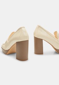 Chaussures à talons hauts en cuir verni beige avec un bout arrondi et un design ajouré. Dotées d'un talon en bois texturé et strié.