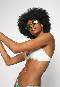 Top de biquíni branco, parte de baixo estampada, óculos de sol verdes e grandes. A modelo tem cabelo preto encaracolado, braços levantados, pele clara com textura suave.