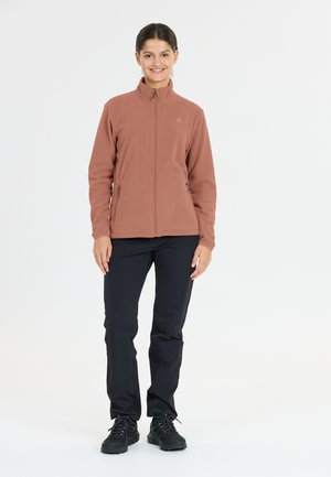 Lachende vrouw staat rechtop, gekleed in een roestkleurige fleecejack met rits, zwarte broek en zwarte wandelschoenen voor een witte achtergrond.