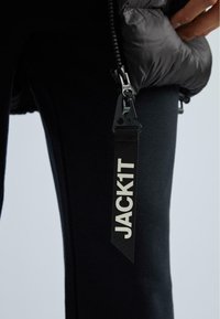 Fekete puff kabát sima textúrával és cipzáros záródással, márkázott hurok címkével, amelyen a "JACK1T" felirat kontrasztos fehér betűkkel olvasható.
