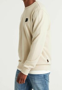 Sweat-shirt beige à manches longues avec encolure ronde. Présente un patch logo noir sur la poitrine et une étiquette sur le côté inférieur. Texture douce.