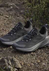 Paire de chaussures de trail Adidas Terrex noires et grises sur un sol rocheux en extérieur avec des plantes vertes en arrière-plan.