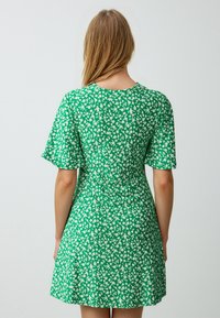 Groene korte jurk met een bloemenprint in het wit, korte uitlopende mouwen en een ronde halslijn. Gemaakt van lichte stof.