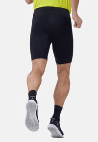 Shorts de sport noirs, longueur genou, présentent une texture lisse avec une taille élastique. Portés avec des chaussettes noires et des chaussures légères.
