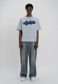 Ανοιχτό μπλε βαμβακερό μπλουζάκι με μαύρο κεντημένο κείμενο που γράφει "Lowlights". Συνδυασμένο με φαρδιά ανοιχτό μπλε τζιν και λευκά αθλητικά παπούτσια.