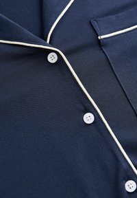 Marineblaue Hemdjacke mit weißen Paspeln, mit Kragen, Brusttasche und drei weißen Knöpfen, aus weichem Stoff gefertigt.