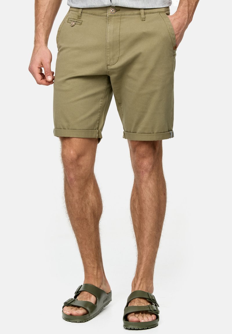 Khakifarbene Baumwollshorts, am Saum gefaltet, mit seitlichen Taschen und Knopfverschluss, kombiniert mit dunkelgrünen Sandalen.