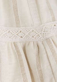 Krämfärgad textil med vertikala linjer av guldtråd, med en virkade spetsmidja och rynkad stil.